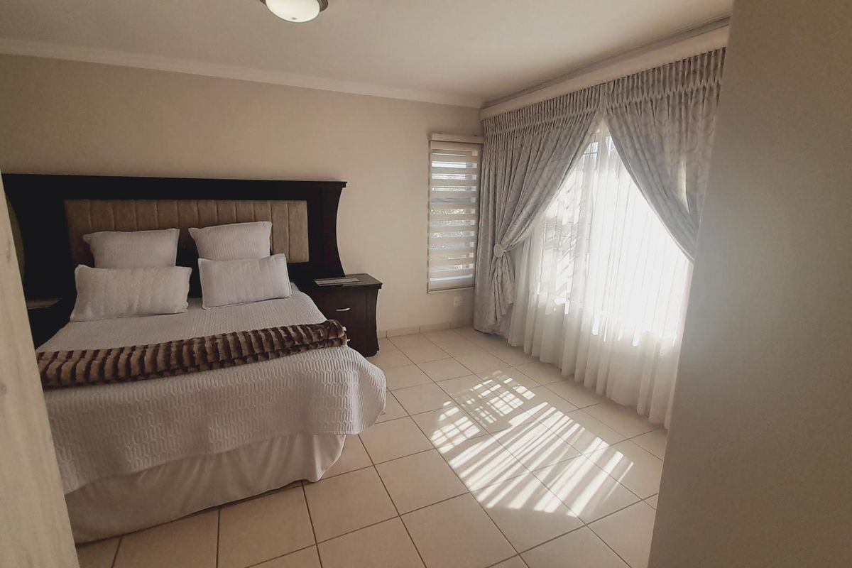 A well-lit bedroom featuring neutral tiled flooring, large windows with fitted curtains, and a built-in headboard structure.