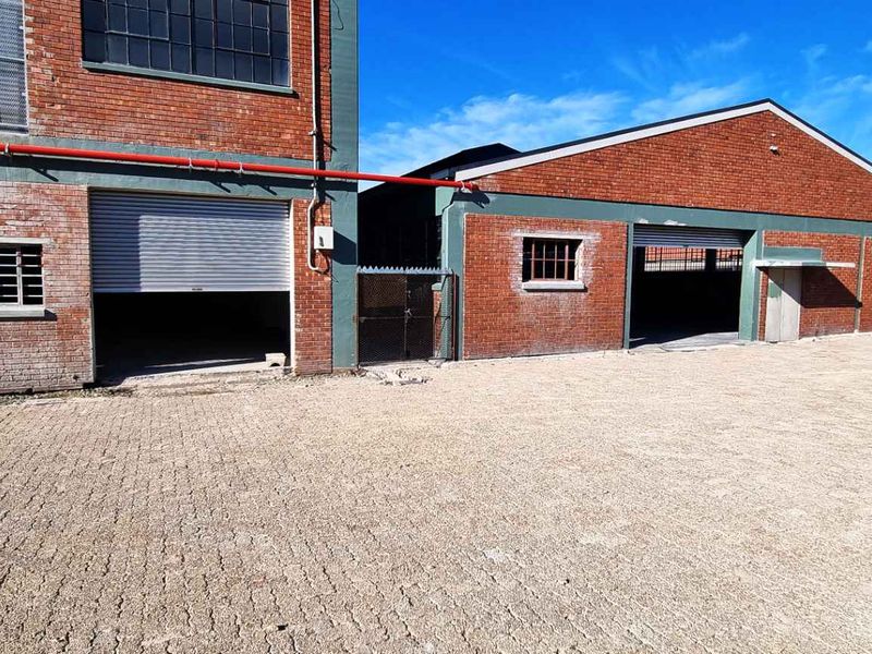 Industrial property exterior featuring brick facade, multiple roller shutter doors, security window bars, and a spacious paved yard area.