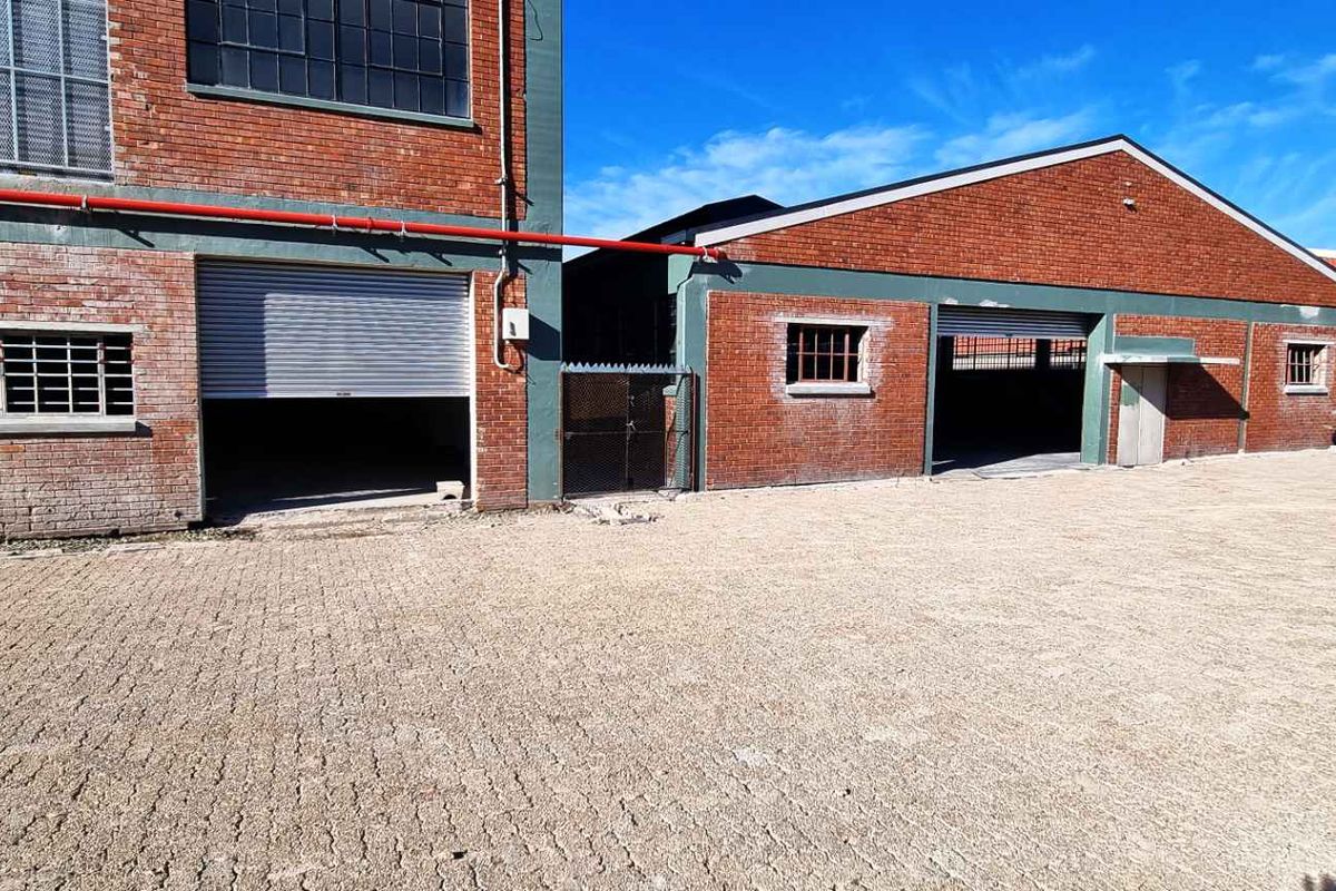 Industrial property exterior featuring brick facade, multiple roller shutter doors, security window bars, and a spacious paved yard area.