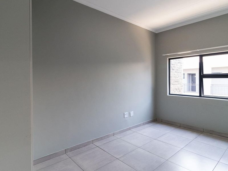 A clean, empty bedroom featuring neutral grey walls, large tiled flooring, and an aluminium-framed window providing natural light.