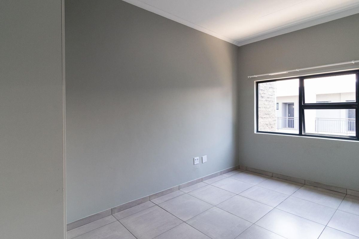 A clean, empty bedroom featuring neutral grey walls, large tiled flooring, and an aluminium-framed window providing natural light.