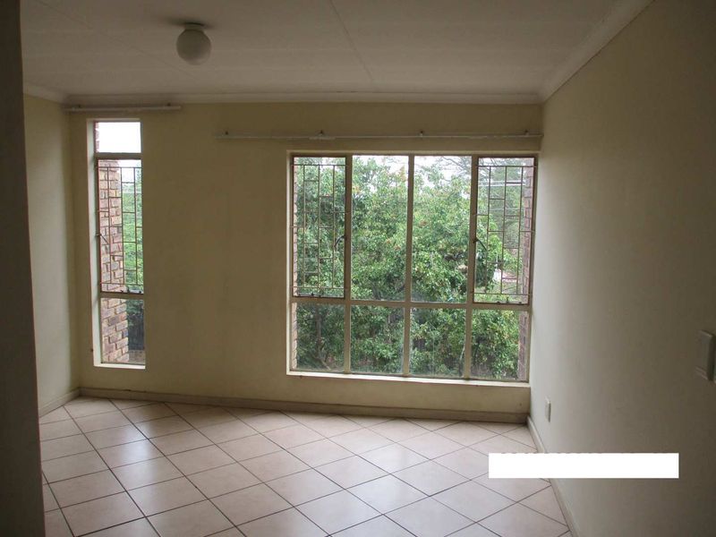 An empty living room featuring tiled flooring and large windows with security bars, offering a view of the surrounding greenery.