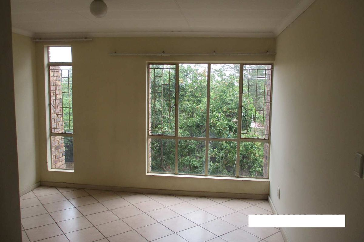 An empty living room featuring tiled flooring and large windows with security bars, offering a view of the surrounding greenery.