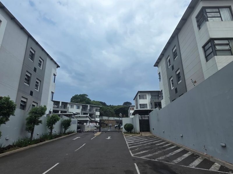 View of the apartment complex entrance featuring a paved driveway, perimeter boundary walls with electric fencing, and multi-story residential building facades.