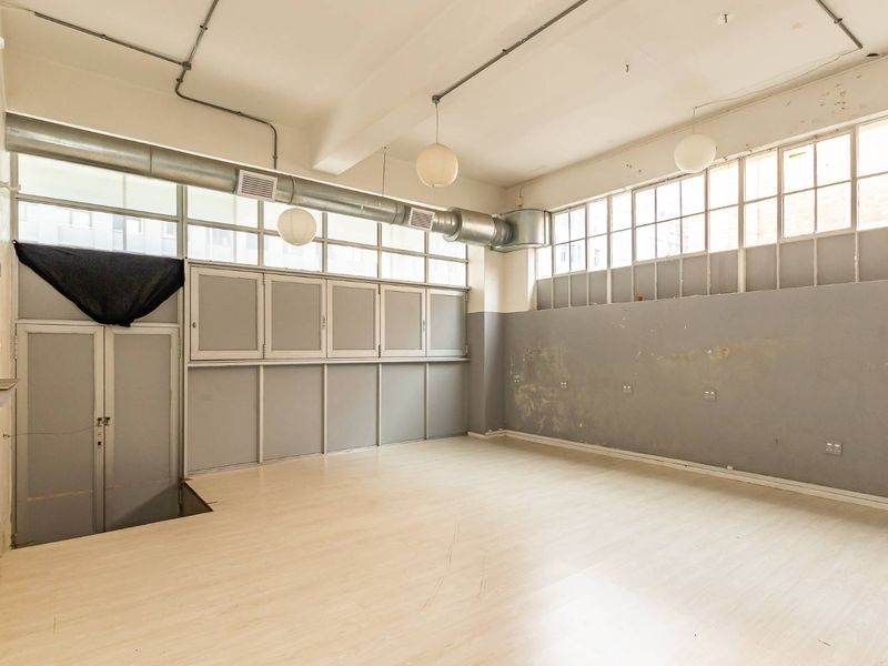 An empty industrial-style open plan space featuring exposed ducting, large windows, built-in cabinetry, and laminate flooring, suitable for a studio or office conversion.
