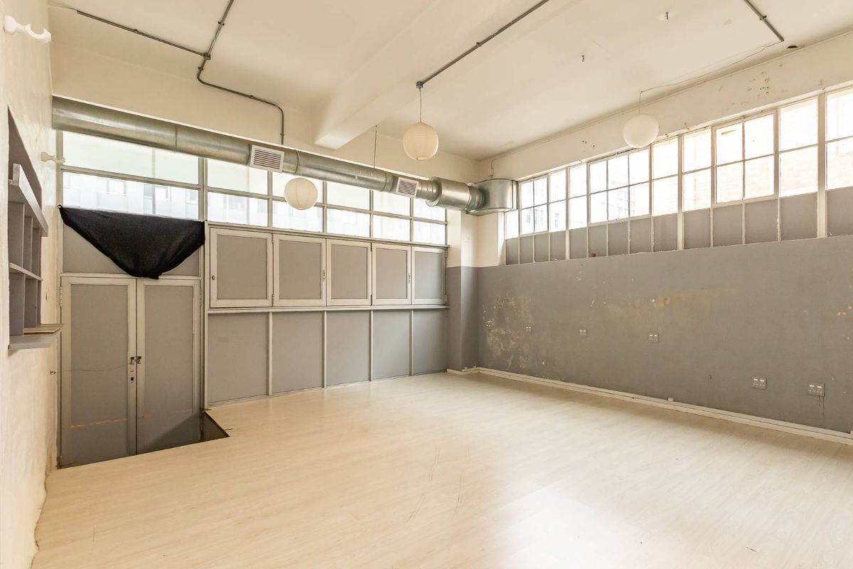 An empty industrial-style open plan space featuring exposed ducting, large windows, built-in cabinetry, and laminate flooring, suitable for a studio or office conversion.