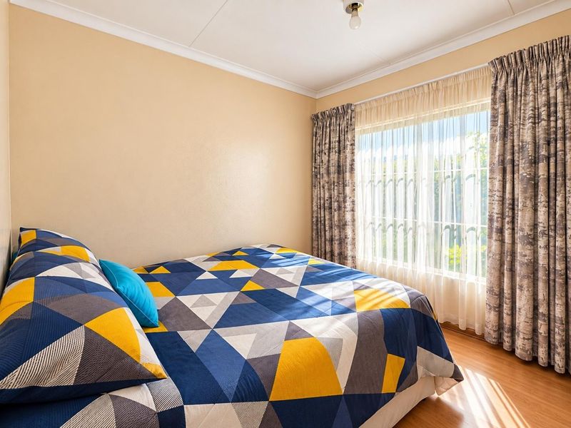 A well-lit bedroom featuring laminate flooring, neutral wall finishes, and large windows fitted with security bars and curtain tracks.