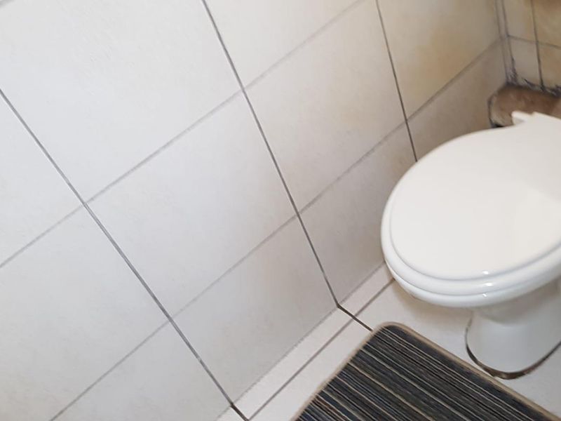 A small bathroom featuring a standard toilet with visible pipework and tiled wall and floor finishes requiring maintenance.