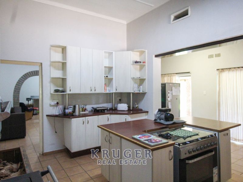 A functional kitchen featuring light-toned cabinetry, laminate countertops, and a central island with a built-in stove, opening into an adjacent living area.