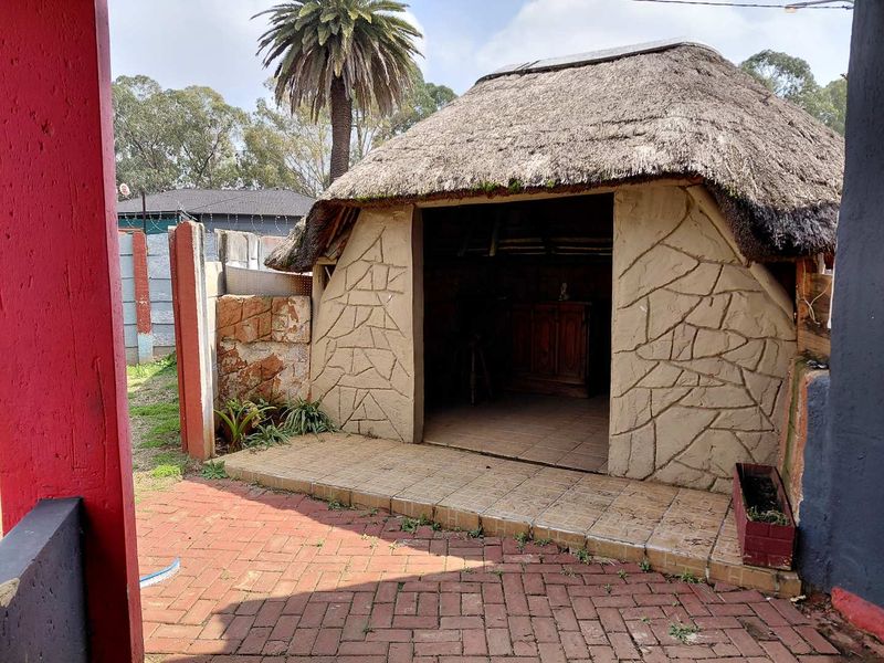 A rustic outdoor entertainment area featuring a traditional thatch roof structure, tiled flooring, and paved patio space within a walled garden setting.