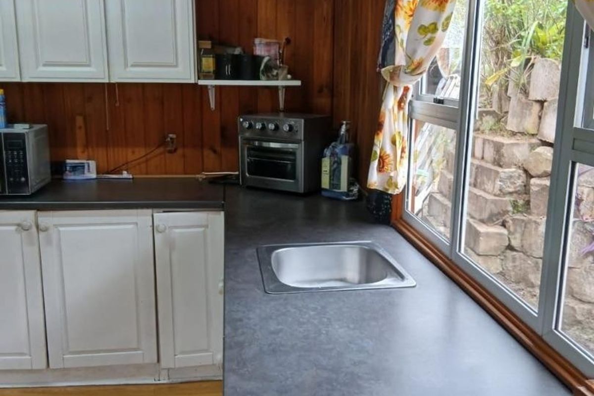 A functional kitchen area featuring timber wall paneling, white cabinetry, dark laminate countertops, and a stainless steel sink with a view of the garden.
