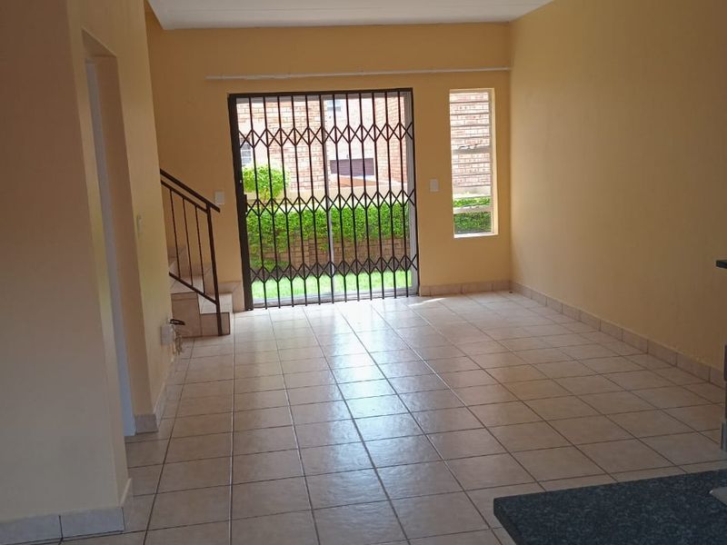 An open-plan living area featuring tiled flooring, a staircase, and a security gate leading to the exterior, providing a bright and functional space.