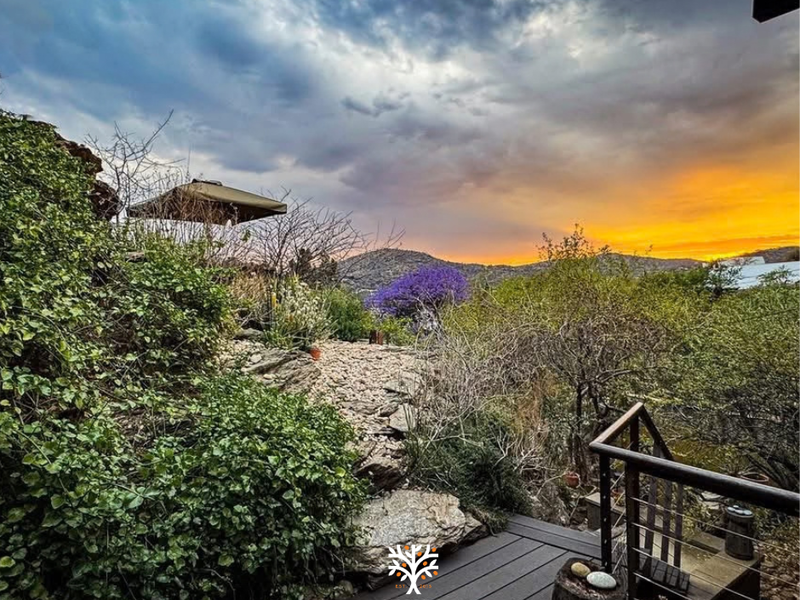 Scenic outdoor view featuring timber decking and metal railing overlooking a landscaped garden and mountain horizon at sunset.