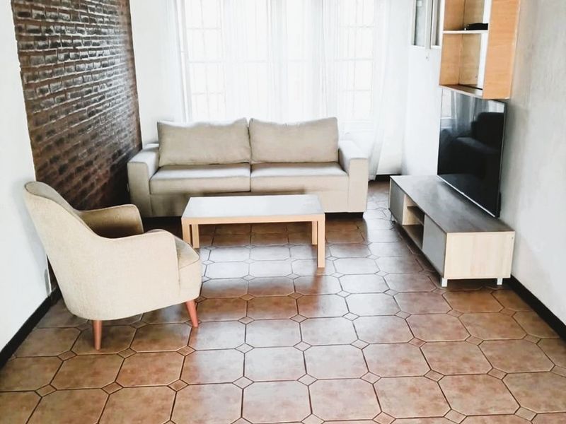 A living room featuring an exposed brick feature wall, timber ceiling, and tiled flooring, providing a rustic aesthetic with natural light from a large window.
