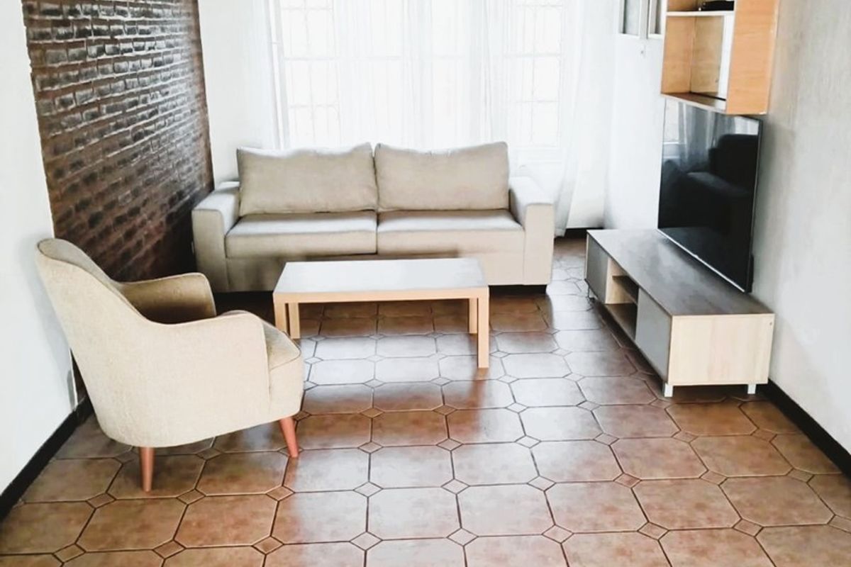 A living room featuring an exposed brick feature wall, timber ceiling, and tiled flooring, providing a rustic aesthetic with natural light from a large window.