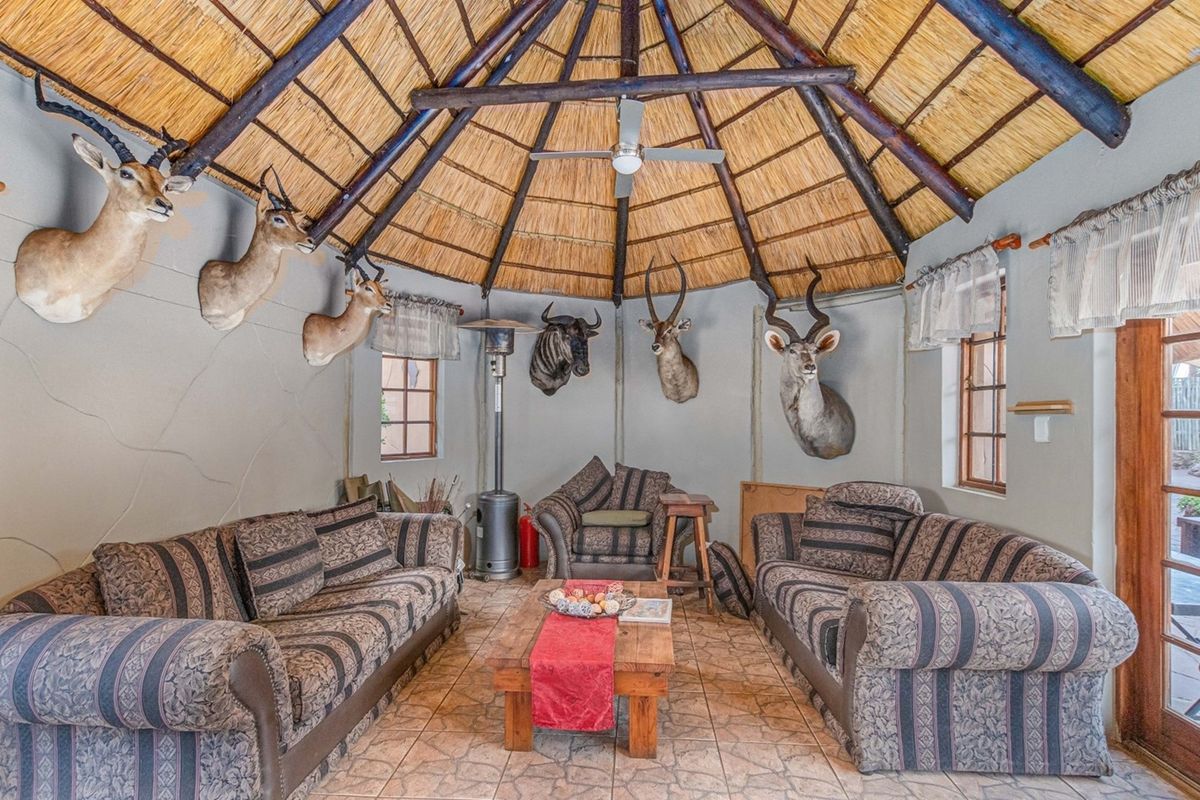 A rustic living area featuring a high thatched roof, exposed timber beams, tiled flooring, and large windows providing natural light.