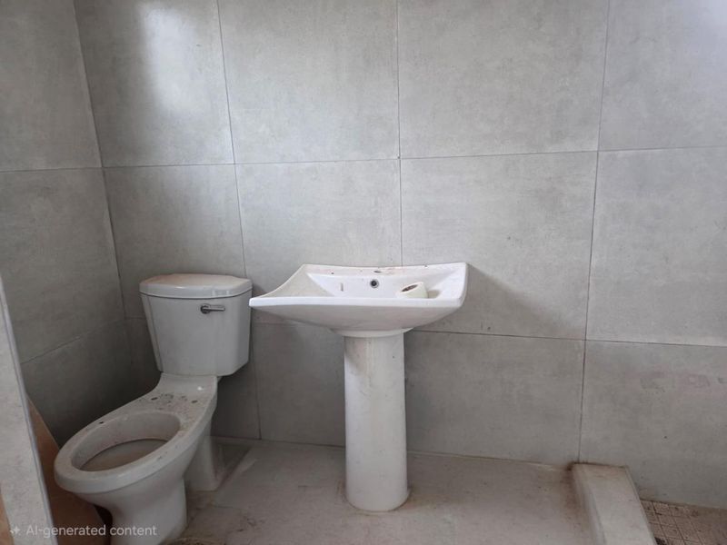A compact bathroom featuring a pedestal basin and toilet with neutral grey wall tiling, requiring cleaning and minor cosmetic updates.
