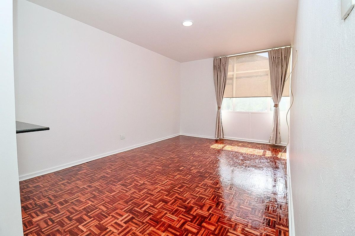 A bright, empty bedroom featuring polished parquet flooring, a built-in wall shelf, and large windows with fitted blinds.