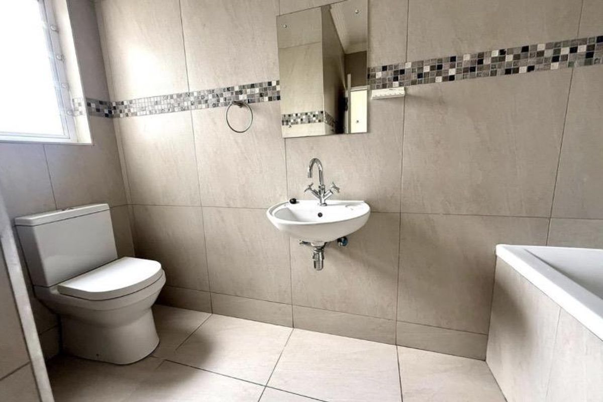 A clean, neutral-toned bathroom featuring a wall-mounted basin, a standard toilet, and a bathtub with full-height wall tiling and a decorative mosaic border.