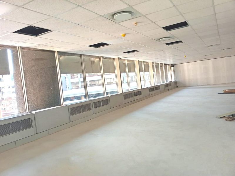 An empty, open-plan office space featuring large windows, perimeter heating units, and a suspended ceiling with some missing tiles.