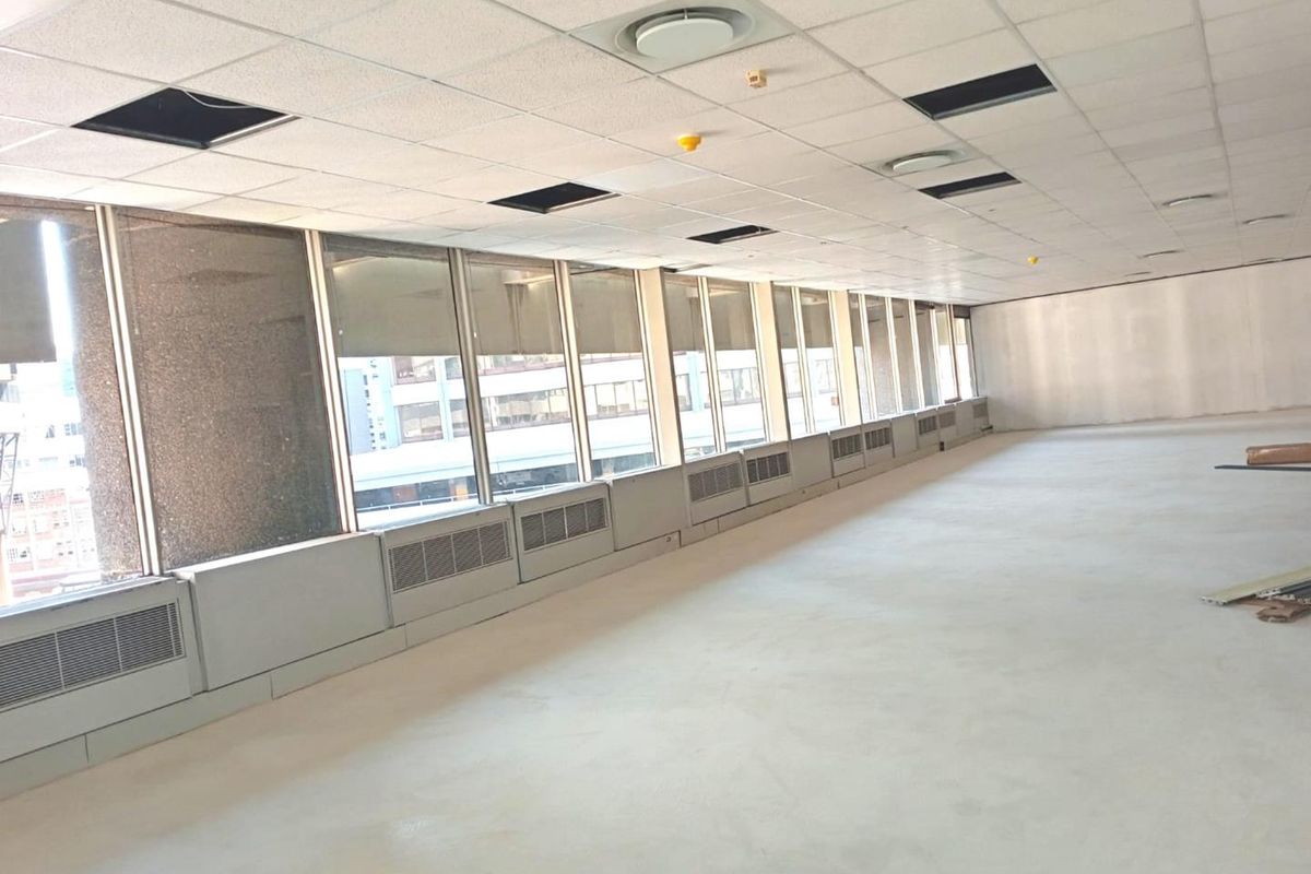 An empty, open-plan office space featuring large windows, perimeter heating units, and a suspended ceiling with some missing tiles.