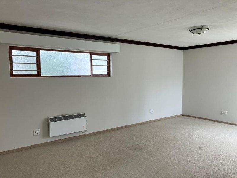 A spacious, empty bedroom featuring neutral walls, carpet flooring, a horizontal sliding window, and a wall-mounted electric heater.