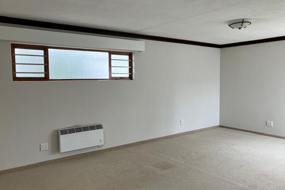 A spacious, empty bedroom featuring neutral walls, carpet flooring, a horizontal sliding window, and a wall-mounted electric heater.