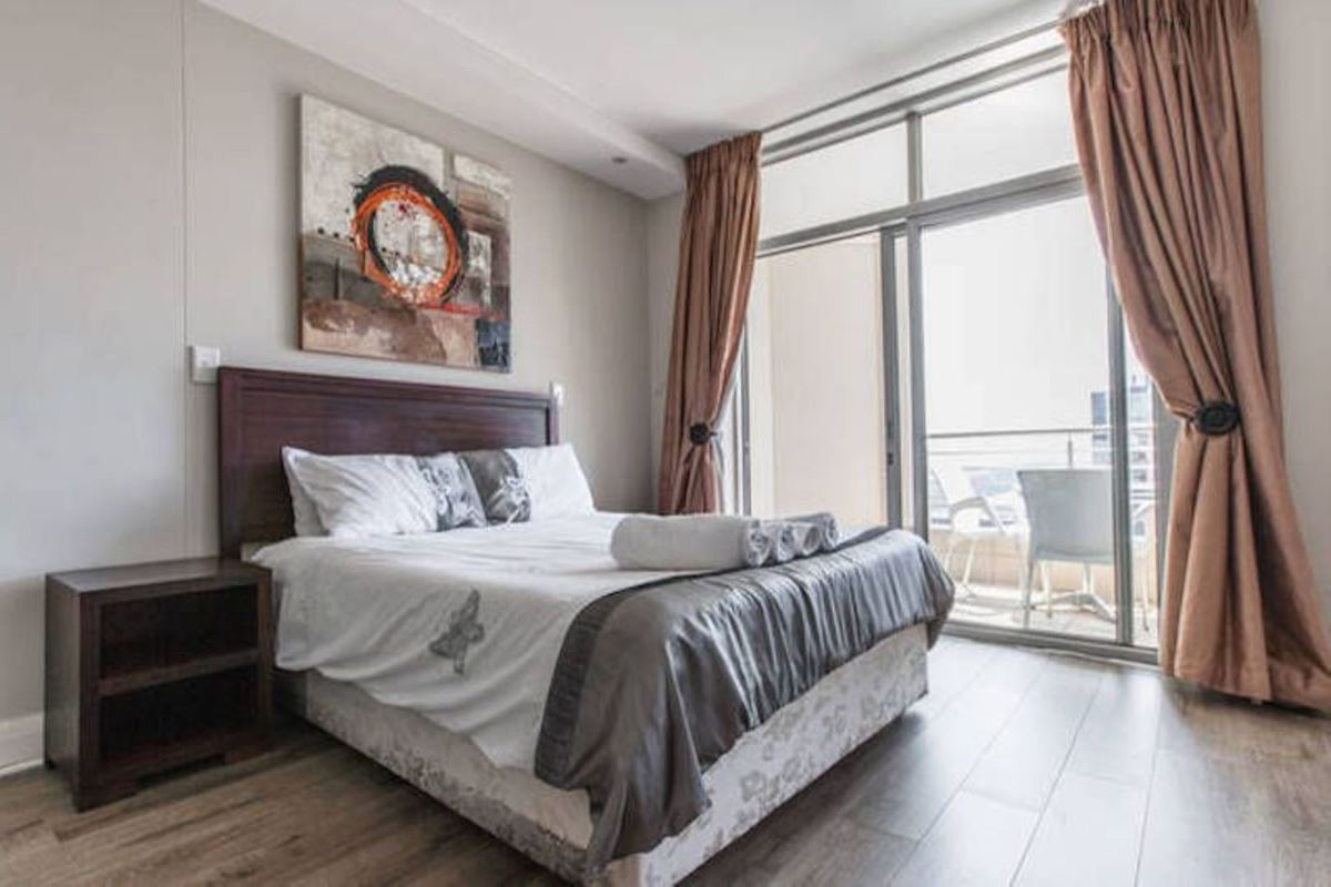 A well-lit bedroom featuring wood-look flooring, large sliding glass doors leading to a private balcony, and neutral wall finishes.