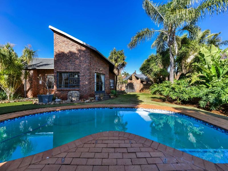 A well-maintained backyard featuring a swimming pool with brick paving, surrounded by a lush garden and a distinctive brick house with a pitched roof.