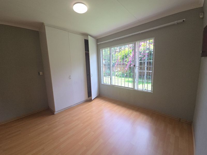 A bright, empty bedroom featuring laminate flooring, built-in cupboards, and large windows with security bars overlooking the garden.