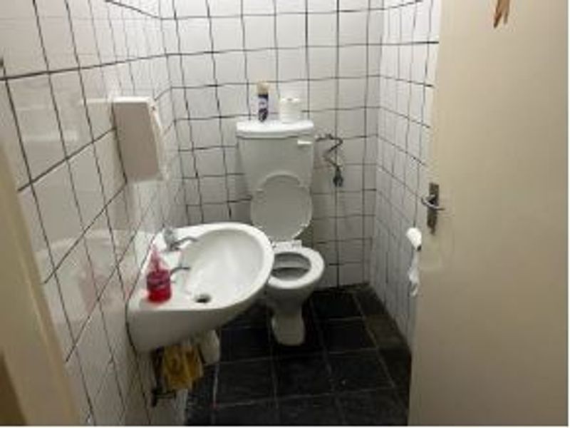 A compact office restroom featuring a wall-mounted basin and toilet with white tiled walls and dark floor tiles, requiring cosmetic updates.