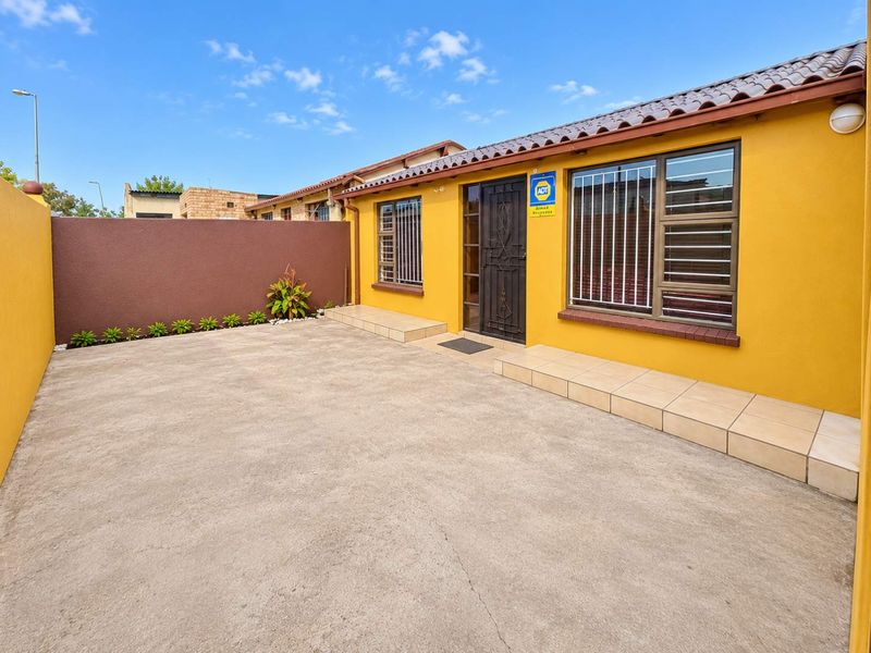 A well-maintained front exterior featuring a concrete driveway, tiled entrance step, and secure window bars, finished with a vibrant yellow facade.