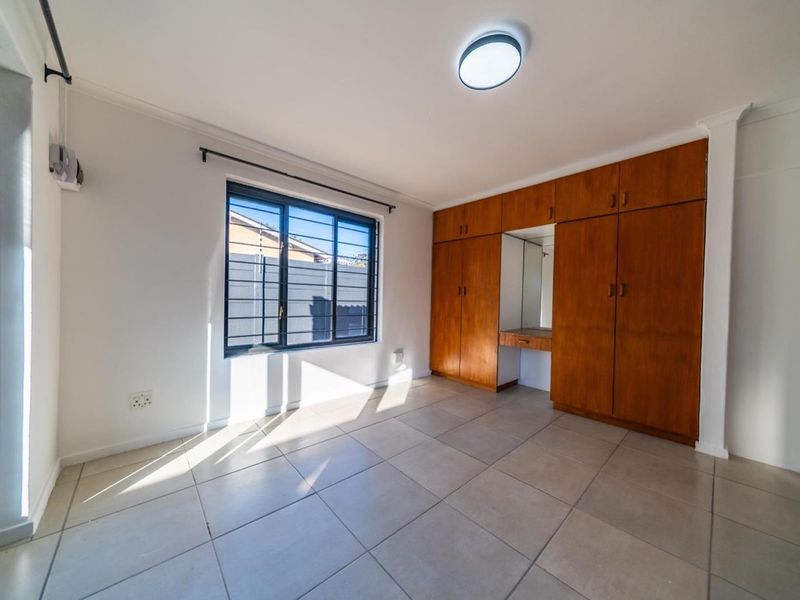 A bright, empty bedroom featuring tiled flooring, built-in wooden cupboards with a vanity section, and a window fitted with security bars.