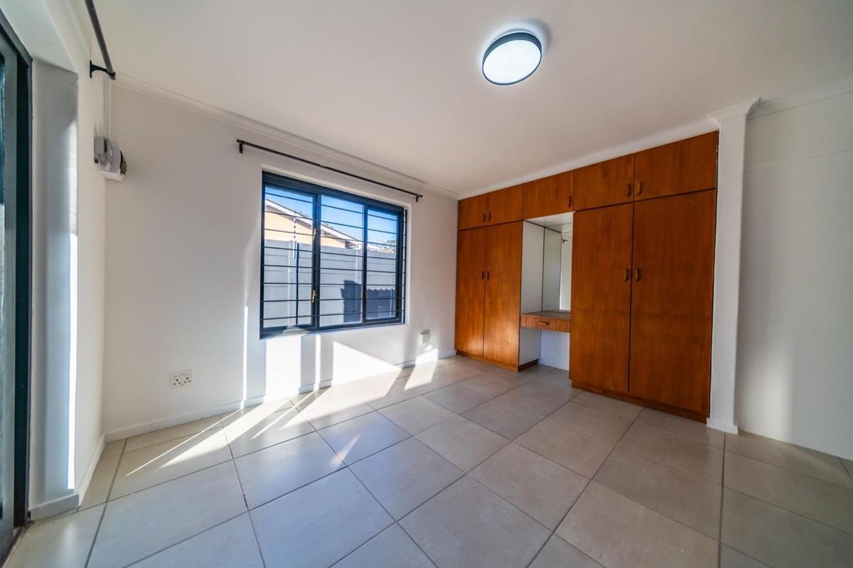 A bright, empty bedroom featuring tiled flooring, built-in wooden cupboards with a vanity section, and a window fitted with security bars.