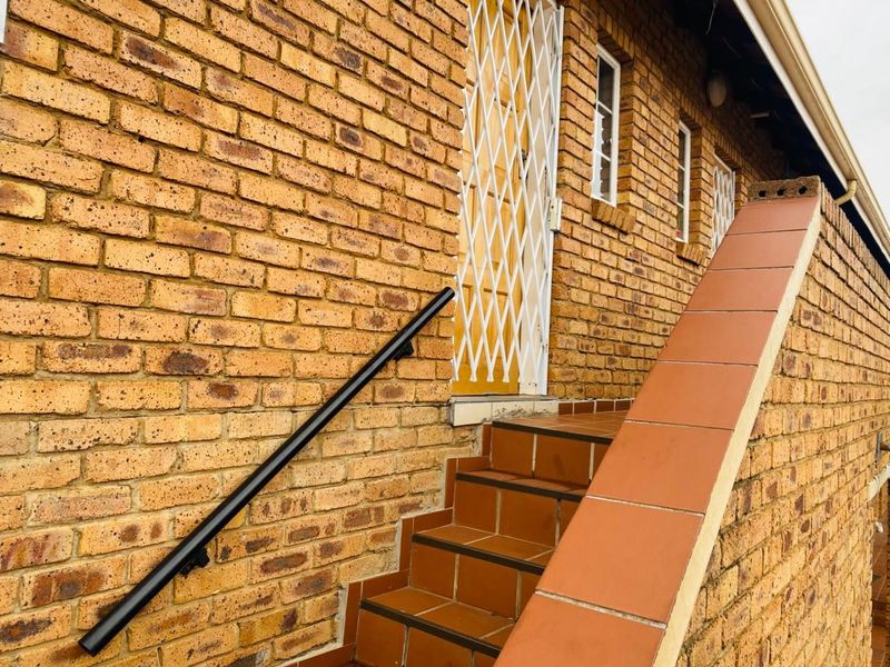 Exterior view of an apartment entrance featuring a brick facade, tiled staircase with a metal handrail, and a security gate.