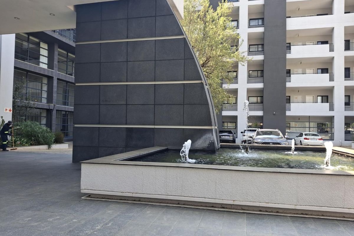 A modern apartment complex courtyard featuring a central water fountain, paved walkways, and multi-story residential building facades with balconies.
