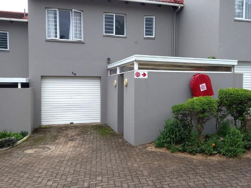Front elevation of a townhouse featuring a paved driveway, single roller garage door, and grey exterior finish with established garden beds.