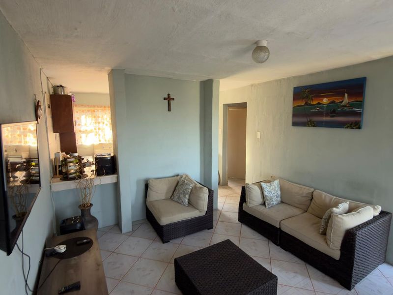 A living room area featuring tiled flooring and direct access to an adjacent kitchen space, showing signs of wear on the wall finishes.