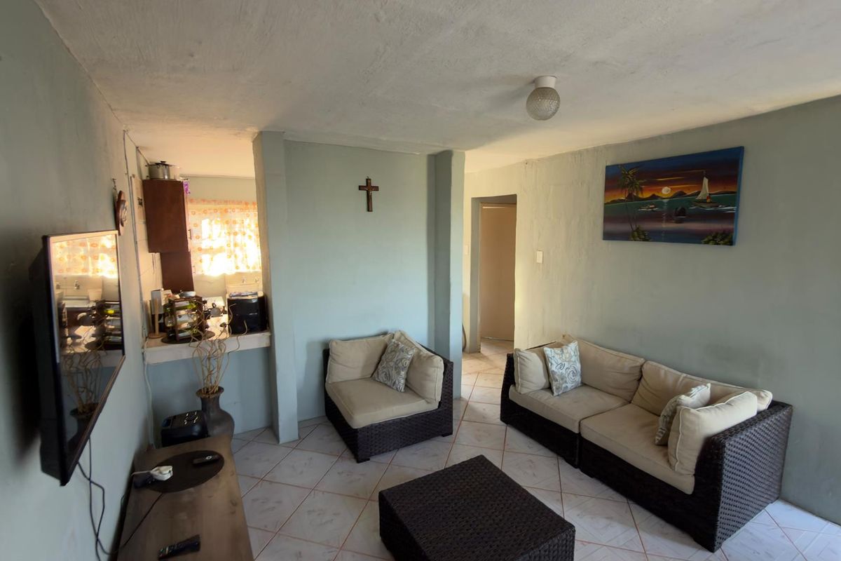 A living room area featuring tiled flooring and direct access to an adjacent kitchen space, showing signs of wear on the wall finishes.