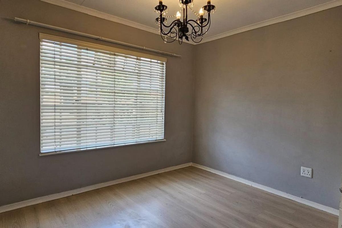 A bright, empty bedroom featuring light wood-look laminate flooring, neutral grey walls, a window with fitted blinds, and a decorative chandelier.