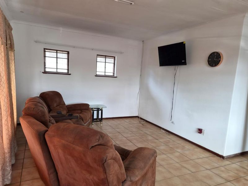 A living room featuring tiled flooring and two small windows, currently furnished with seating and a wall-mounted television.