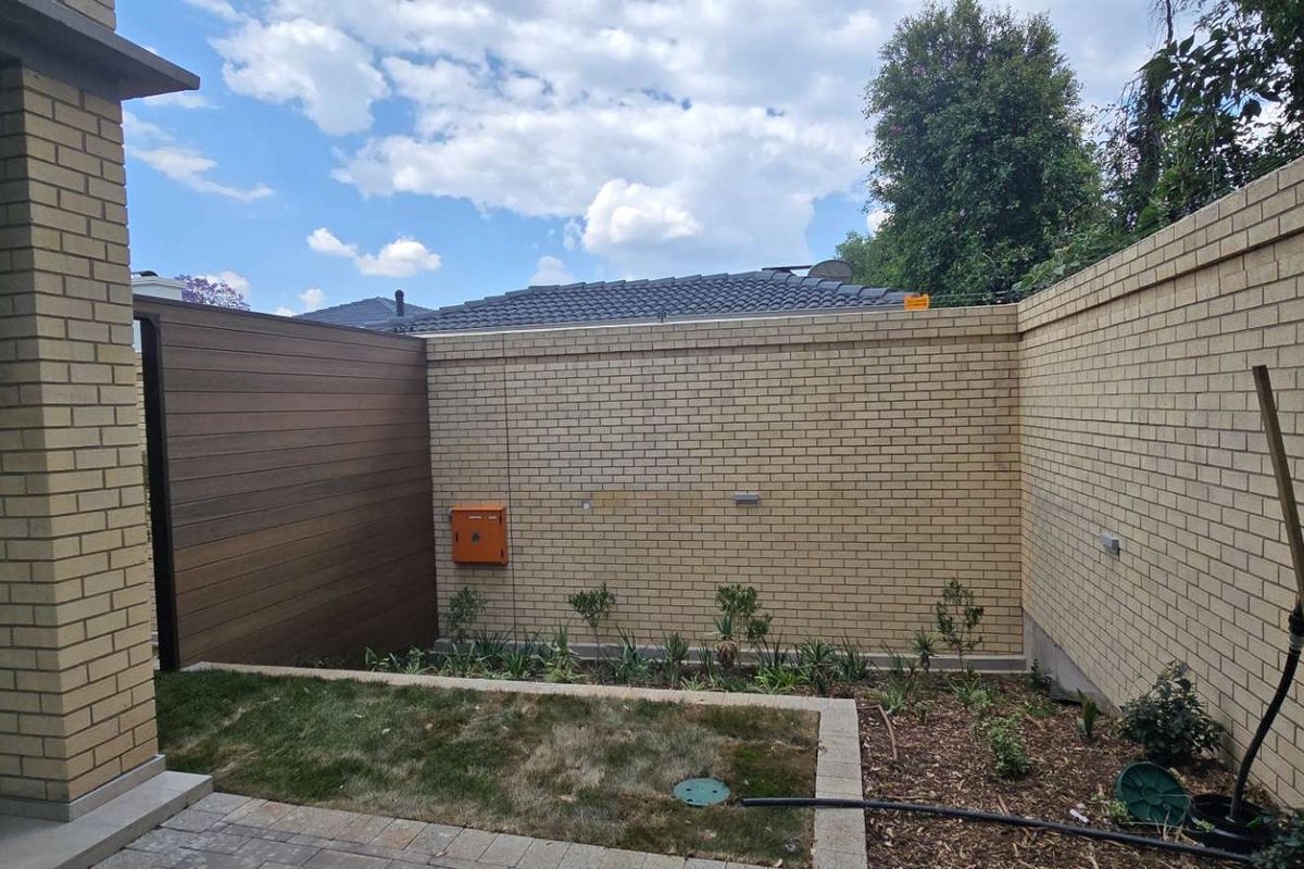 A private rear courtyard featuring a brick boundary wall, timber privacy screen, paved patio area, and a small landscaped garden bed.