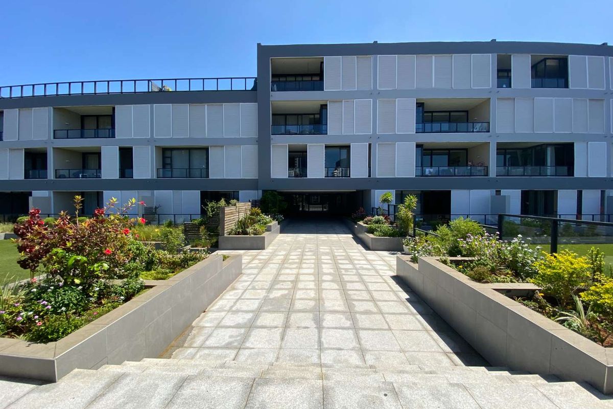 A well-maintained modern apartment complex featuring a paved walkway, landscaped garden beds, and private balconies with glass balustrades.