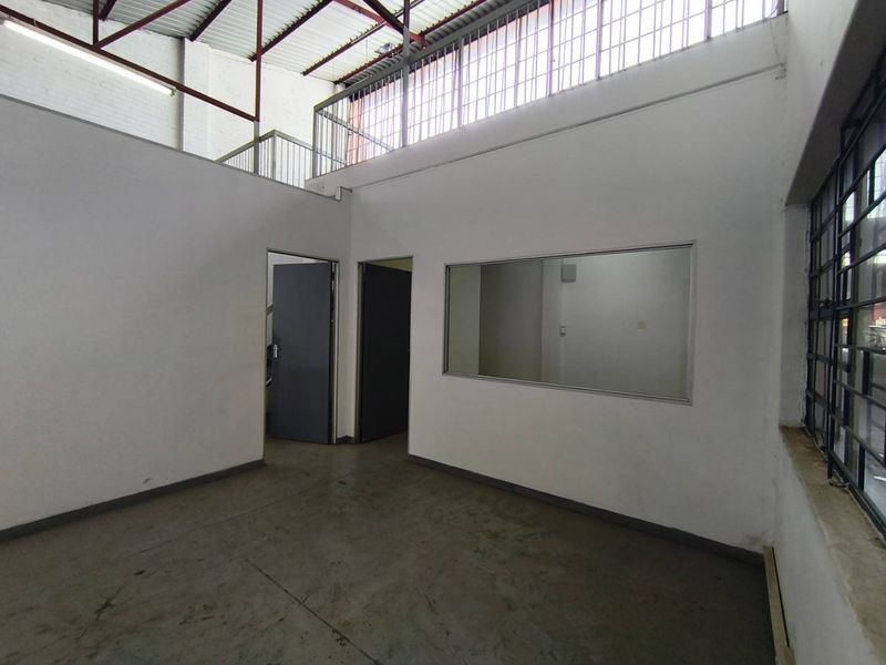 Industrial office space featuring a mezzanine level, exposed roof trusses, concrete flooring, and an internal viewing window, offering a functional workspace layout.