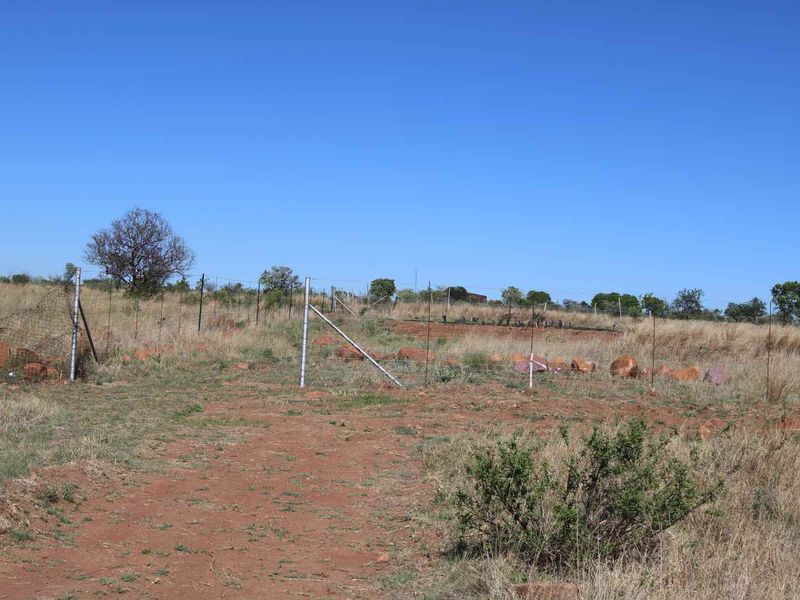 An expansive, undeveloped plot of land featuring natural dry grass, scattered rocks, and perimeter wire fencing under a clear blue sky.