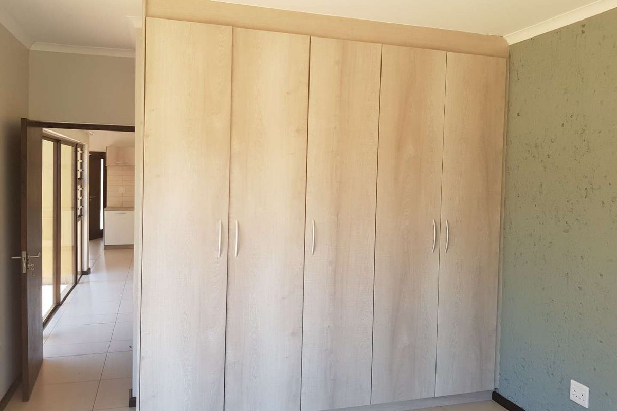 A bedroom featuring light wood-finish built-in cupboards, tiled flooring, and a textured accent wall, with a view into the adjacent hallway.