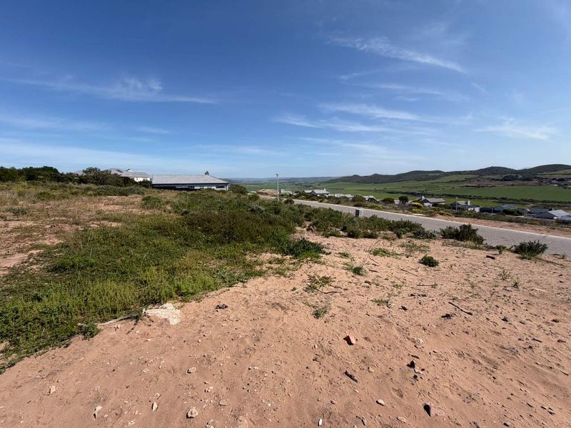 A wide-angle view of a vacant residential plot featuring natural vegetation and a paved road with surrounding landscape views.