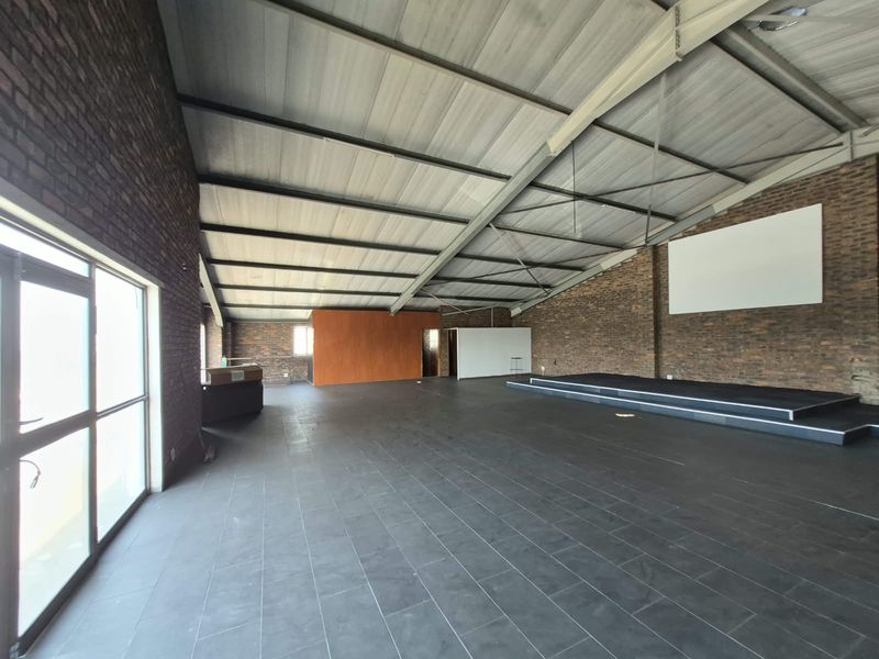 Spacious open-plan office area featuring exposed brick walls, tiled flooring, a raised stage platform, and high pitched ceilings with ample natural light.