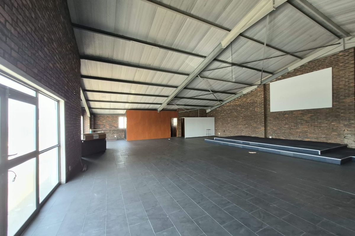 Spacious open-plan office area featuring exposed brick walls, tiled flooring, a raised stage platform, and high pitched ceilings with ample natural light.