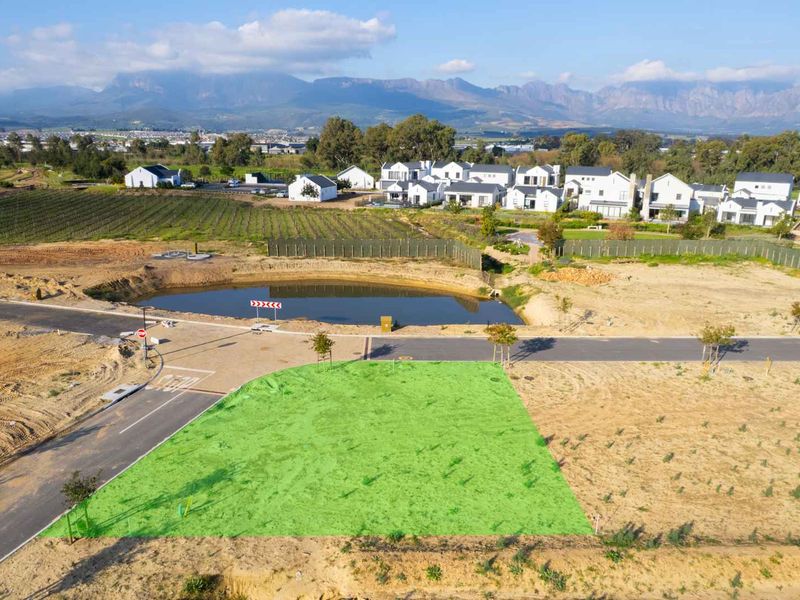 Aerial view of a vacant residential plot in a developed estate, featuring road access and proximity to a scenic retention pond and mountain views.
